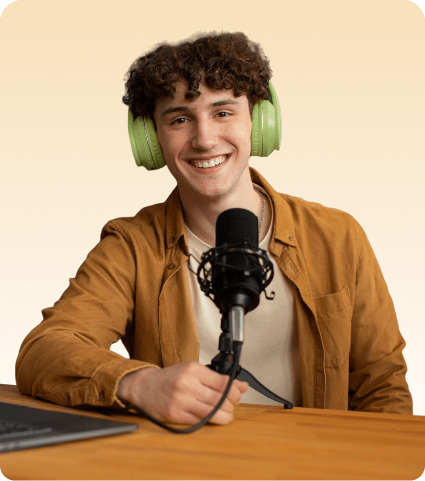 A smiling young man wearing bright green over-ear headphones sits at a wooden desk in front of a professional microphone, with a laptop beside him against a soft beige background.